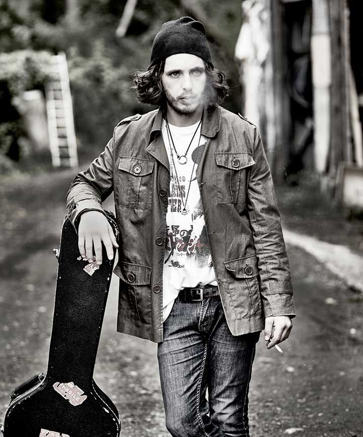 Photo de Jess Creatchman debout, appuyé sur un étui de guitare, en train de fumer une cigarette