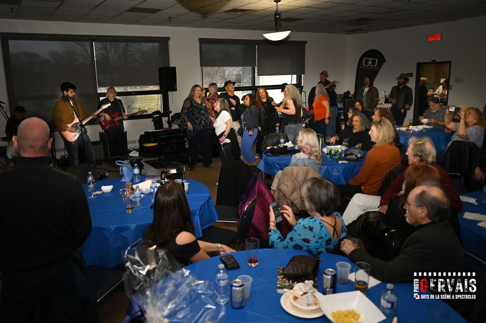 Crowd sitting at round tables with blue tablecloths, smiling and watching the band play