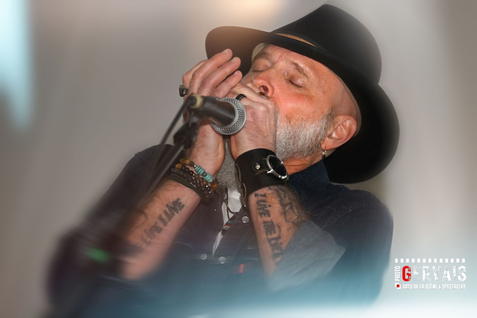 A man in a black hat, black shirt with both arms tattooed playing a harmonica