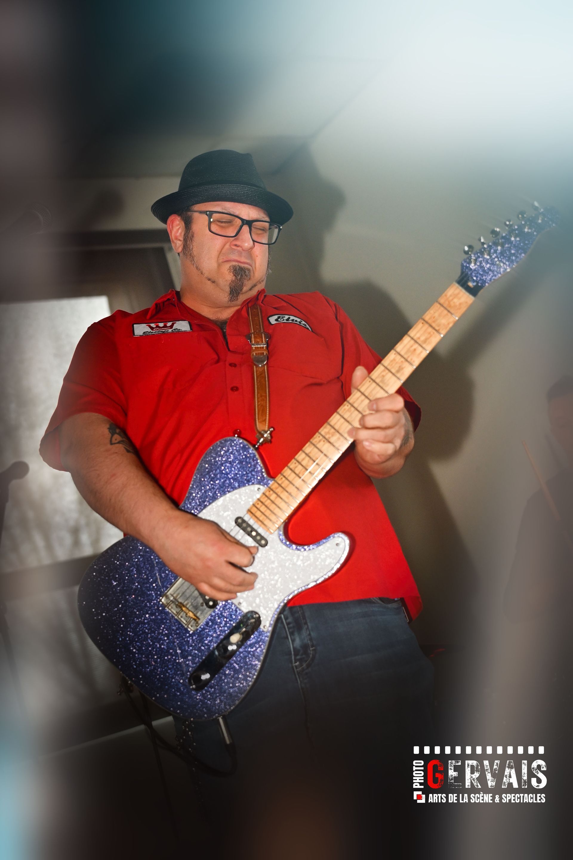 A man in a red shirt, black hat playing a retro looking guitar