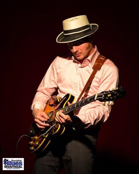 Jordan Officer on stage, sporting a black and white blues hat that coordinates with his shirt, looking smart as he plays his guitar.