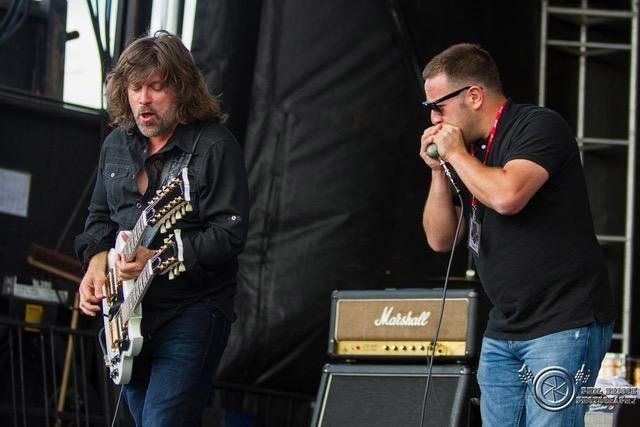 Paul Deslauriers sporting his hair long, playing guitar on stage with a harmonica player.