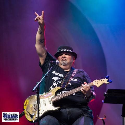 Popa Chubby on stage, sitting down with guitar and holding arm up with a 2 finger hand gesture.