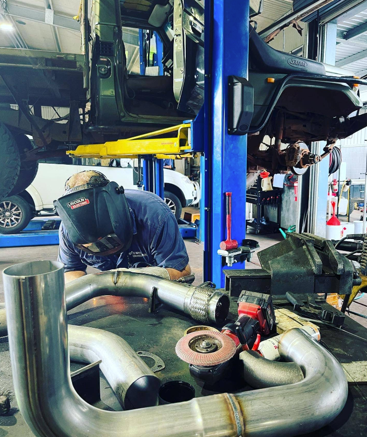 Welder In A Shop Working On Car Exhaust System Parts. Car Elevated, Wearing Welding Helmet, Blue Shirt — Atherton Mechanical Services in Atherton, QLD