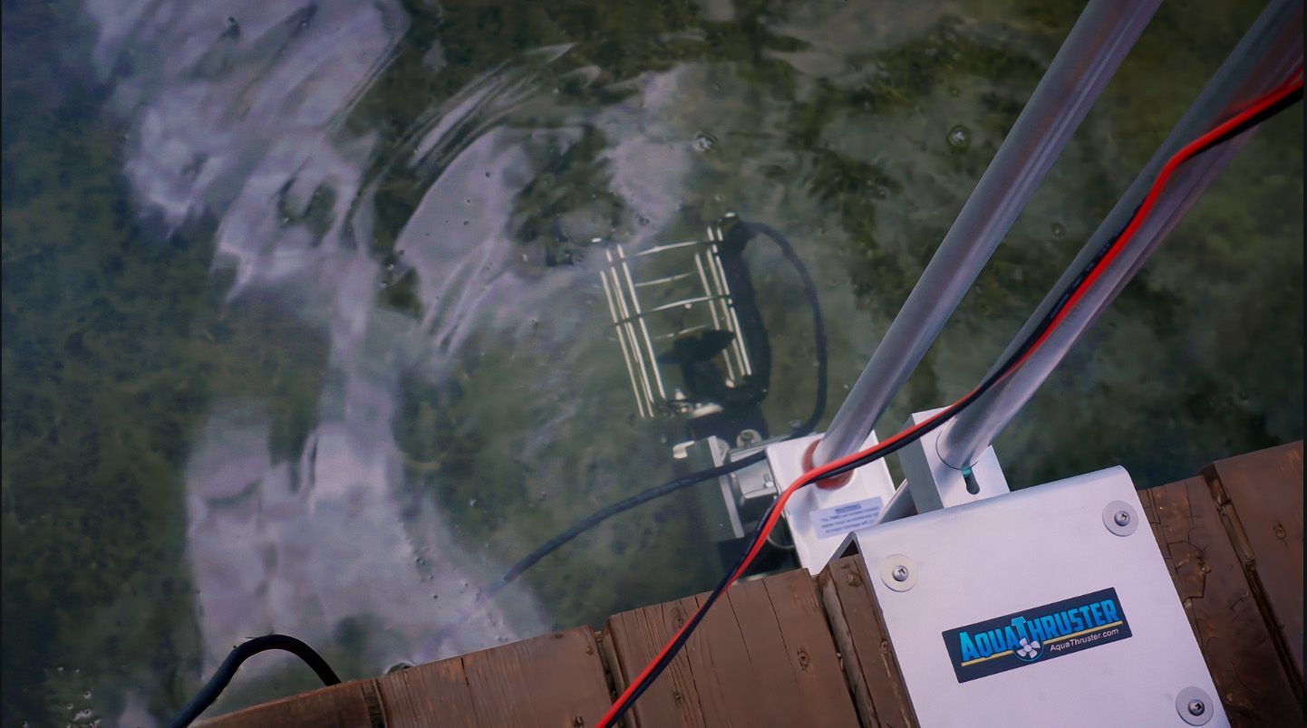 Device submerged in water, connected to a white box with wires on a wooden dock cleaning algae