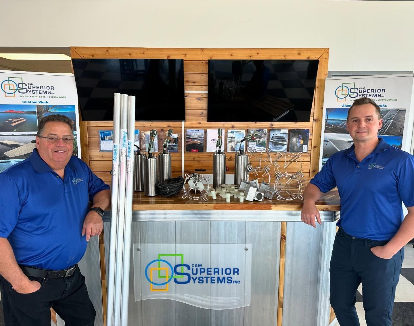 Two men in blue shirts pose with a display of metal parts. A logo for 