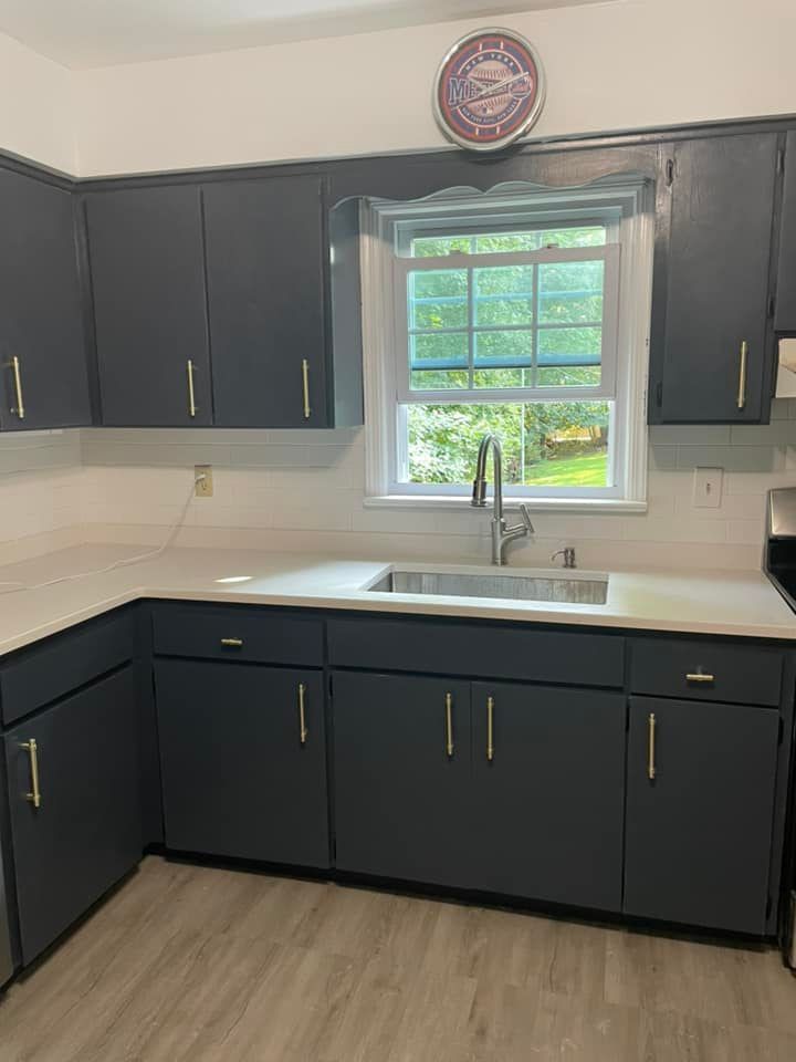 A kitchen featuring dark blue cabinets with gold handles, a white countertop, a silver sink, and a window with a view.