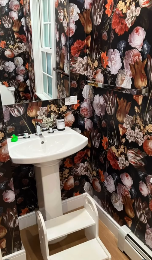 A white pedestal sink with a two-step stool in front, set against a dark floral wallpaper background.