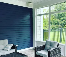 A sunroom with a dark blue shiplap wall, light flooring, two wicker chairs with grey cushions, and large windows.