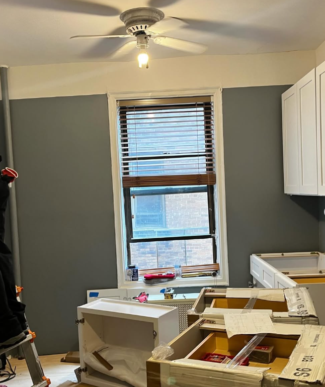 A kitchen under renovation with gray walls, white cabinets, and a window with wooden blinds under a ceiling fan.