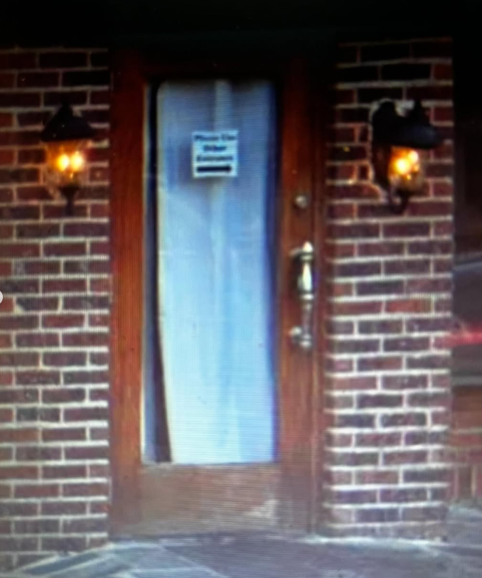 A brown wooden door centered in a brick wall, flanked by two outdoor lanterns, with a sign directing to another entrance.