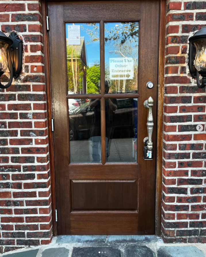 A dark wood door set in a red brick wall, with a sign in the glass pointing right that reads 