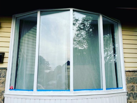 A white bay window framed by tan siding and a stone foundation, with blue painter's tape along the base of the glass.