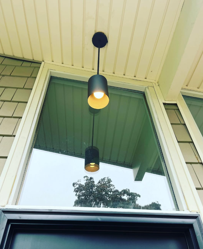 A black, cylindrical pendant light hanging from a porch ceiling, with its reflection visible in a tall window below.