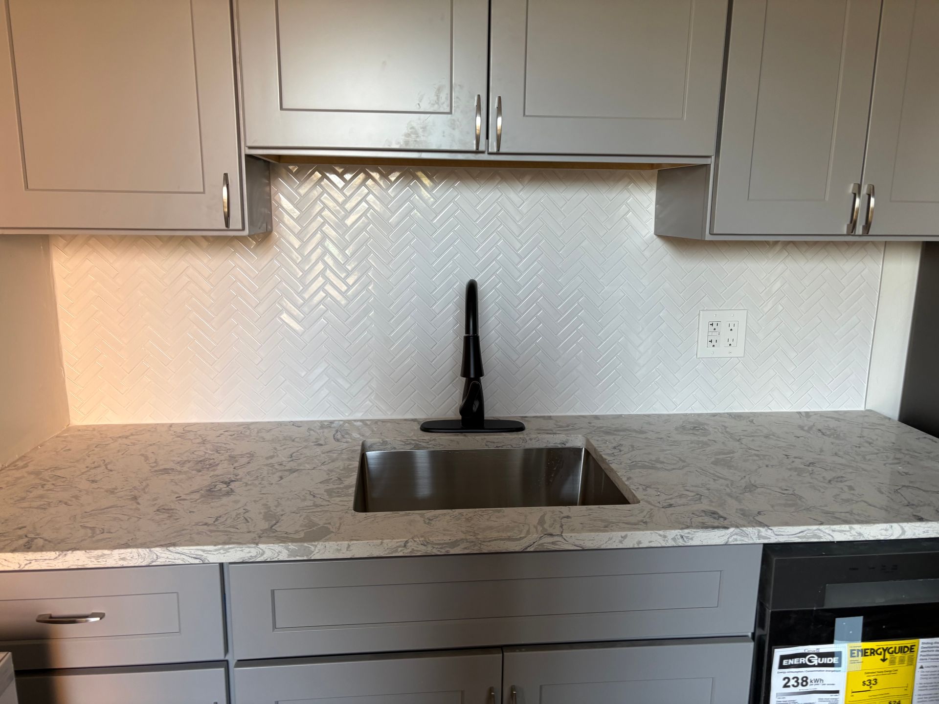 A kitchen sink area featuring grey cabinets, a speckled light-grey countertop, a black faucet, and white herringbone tiles.