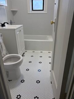 A small, bright bathroom with white walls, a white vanity, a toilet, a bathtub, and white-and-black floral-patterned tile.