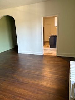 An empty room with dark wood floors, white walls, an arched entryway, and a doorway leading to a kitchen.