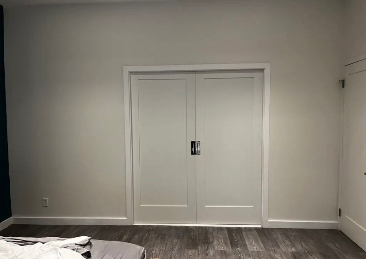 A pair of white, shaker-style double doors centered on a light gray wall above wood-look flooring.