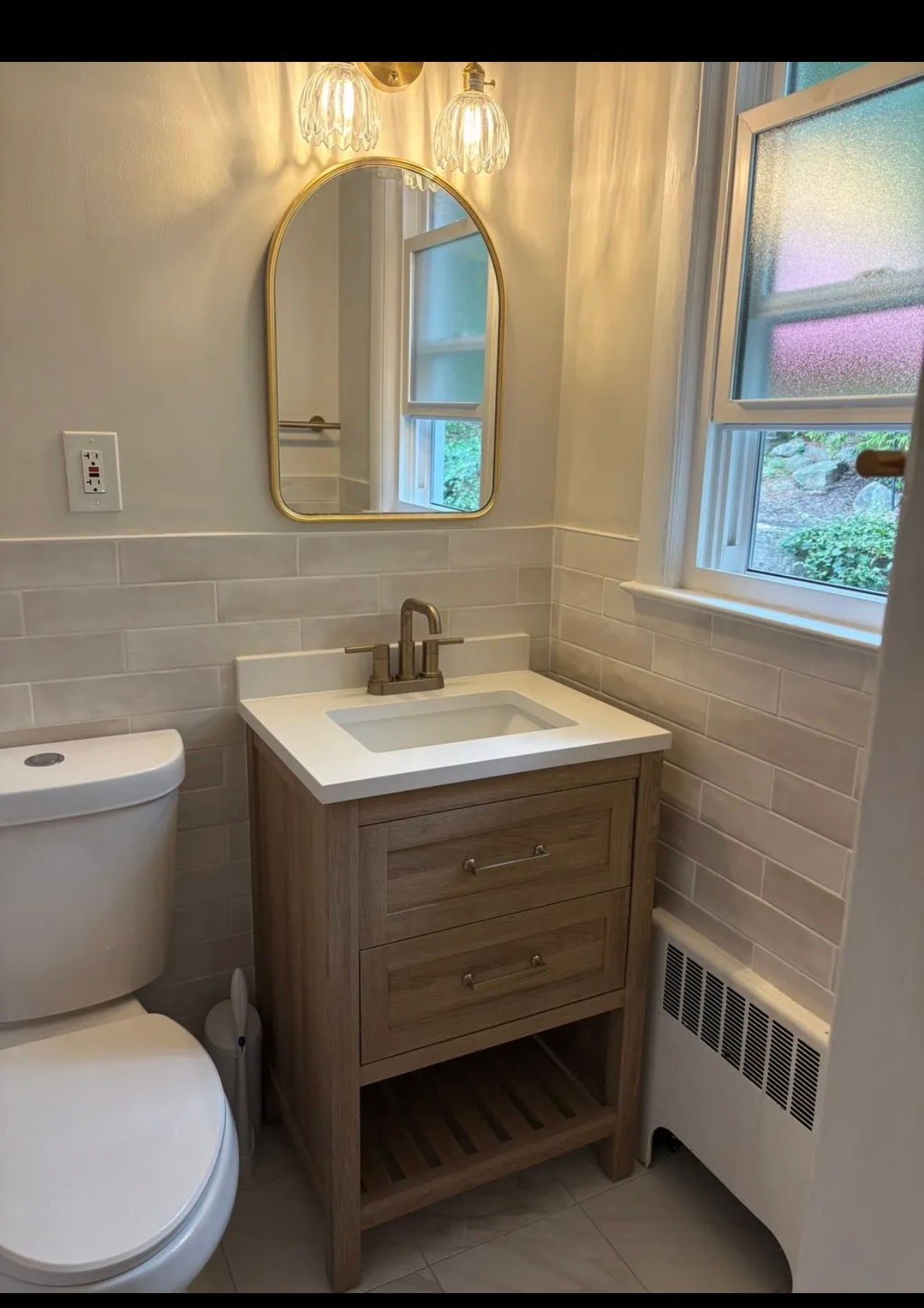 A small bathroom features a light wood vanity, white tiled walls, an arched gold-rimmed mirror, and a toilet.