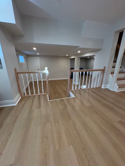 A room with light wood-tone flooring, white walls, and a wooden staircase railing leading to a lower level.
