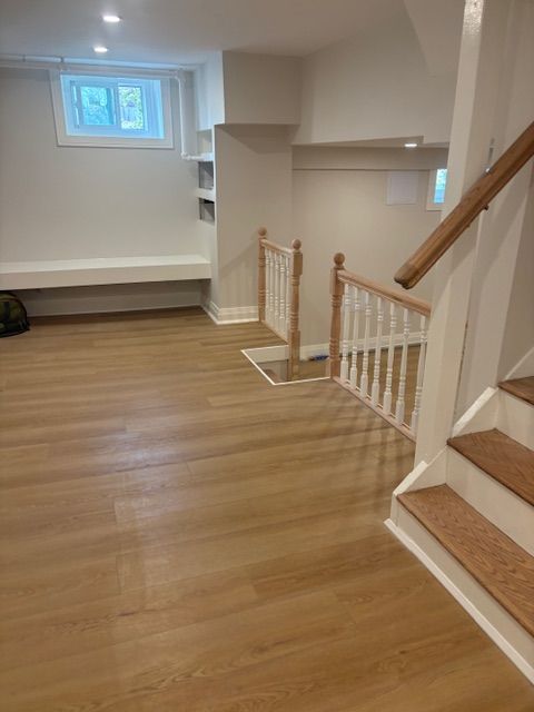A renovated basement with light wood flooring, white walls, a built-in window seat, and wooden stairs with a white railing.