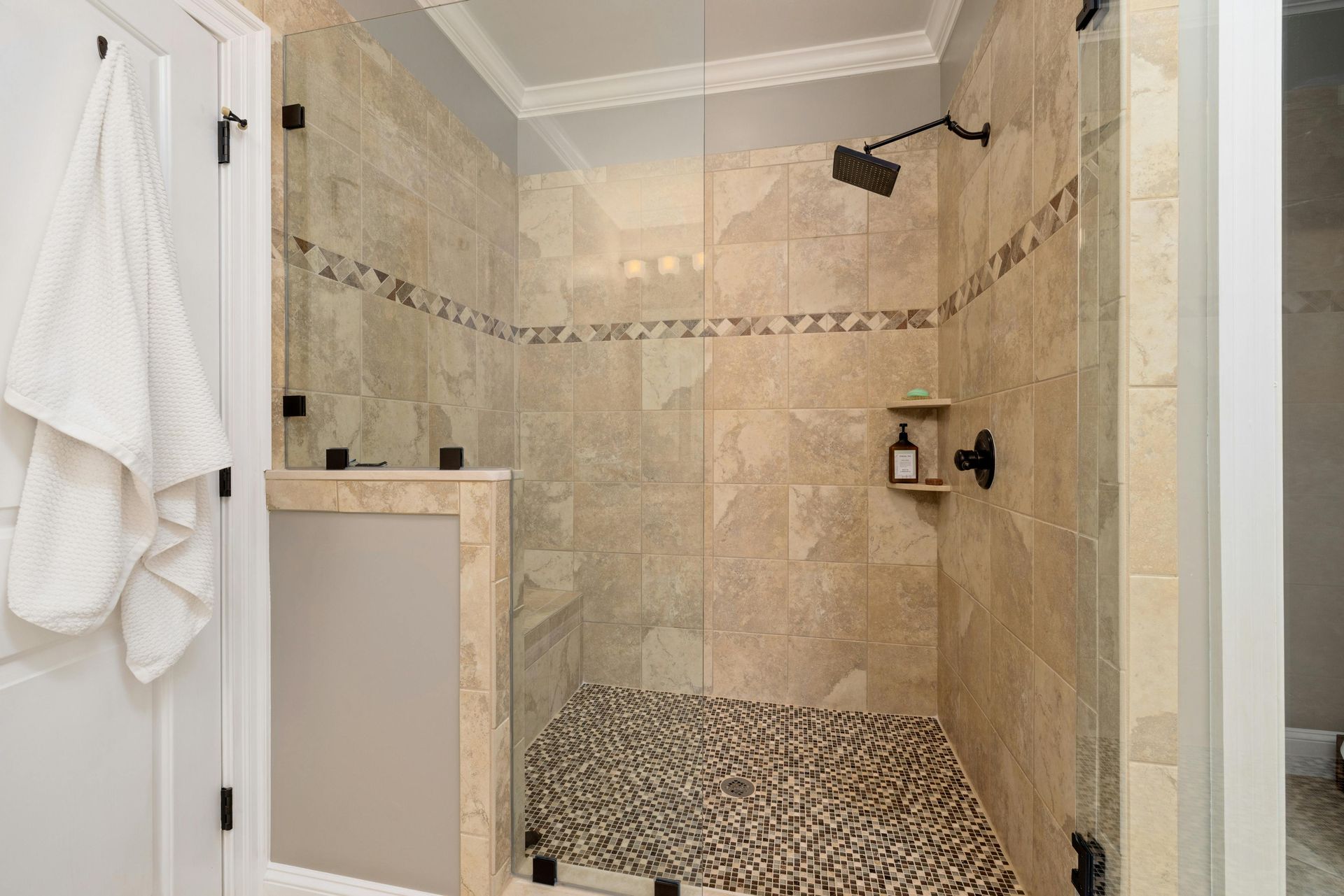 A walk-in shower with tan tiled walls, a decorative stone border, a pebble floor, and dark hardware.