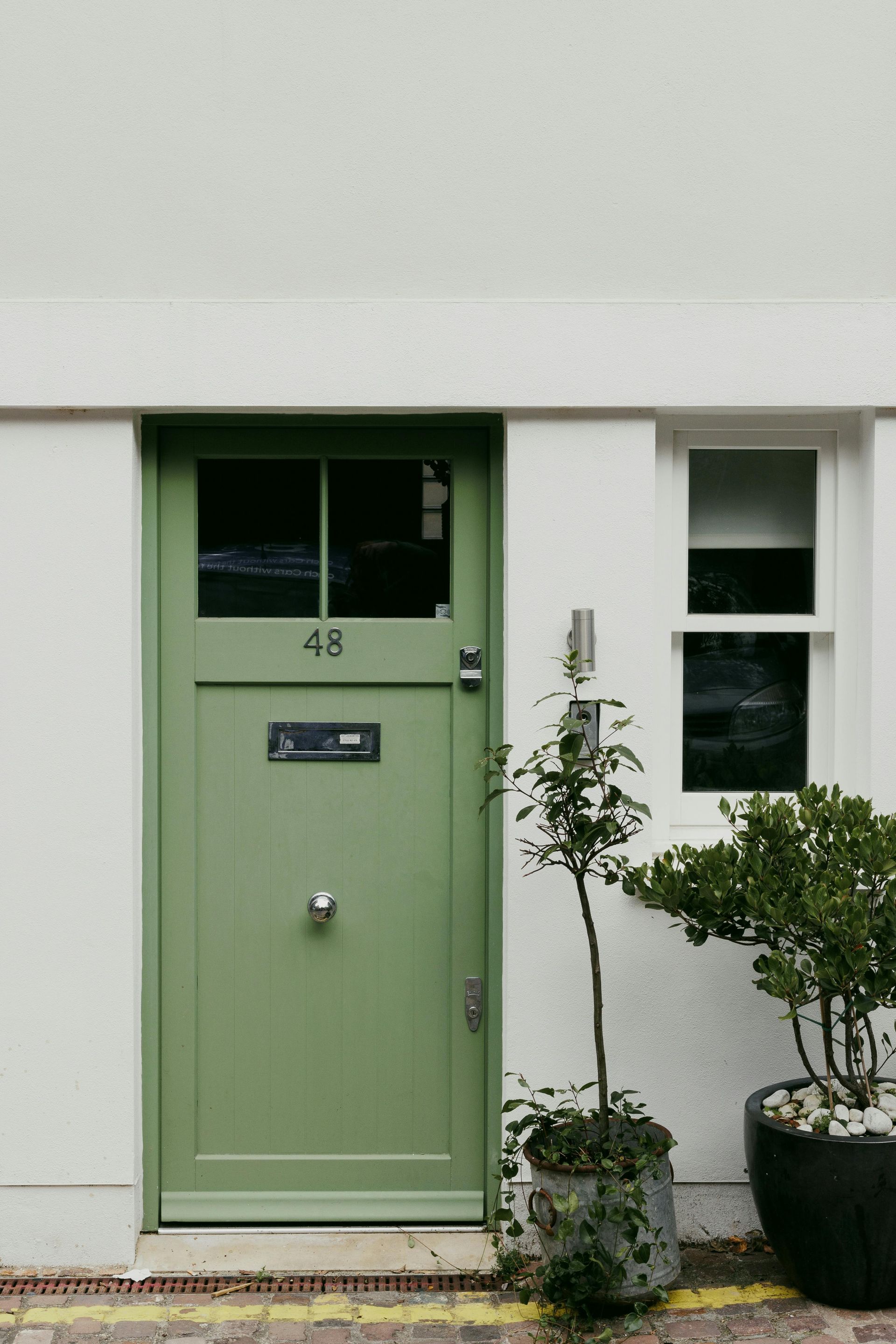 A sage green wooden front door numbered 48, set in a white wall next to a window and two potted plants.