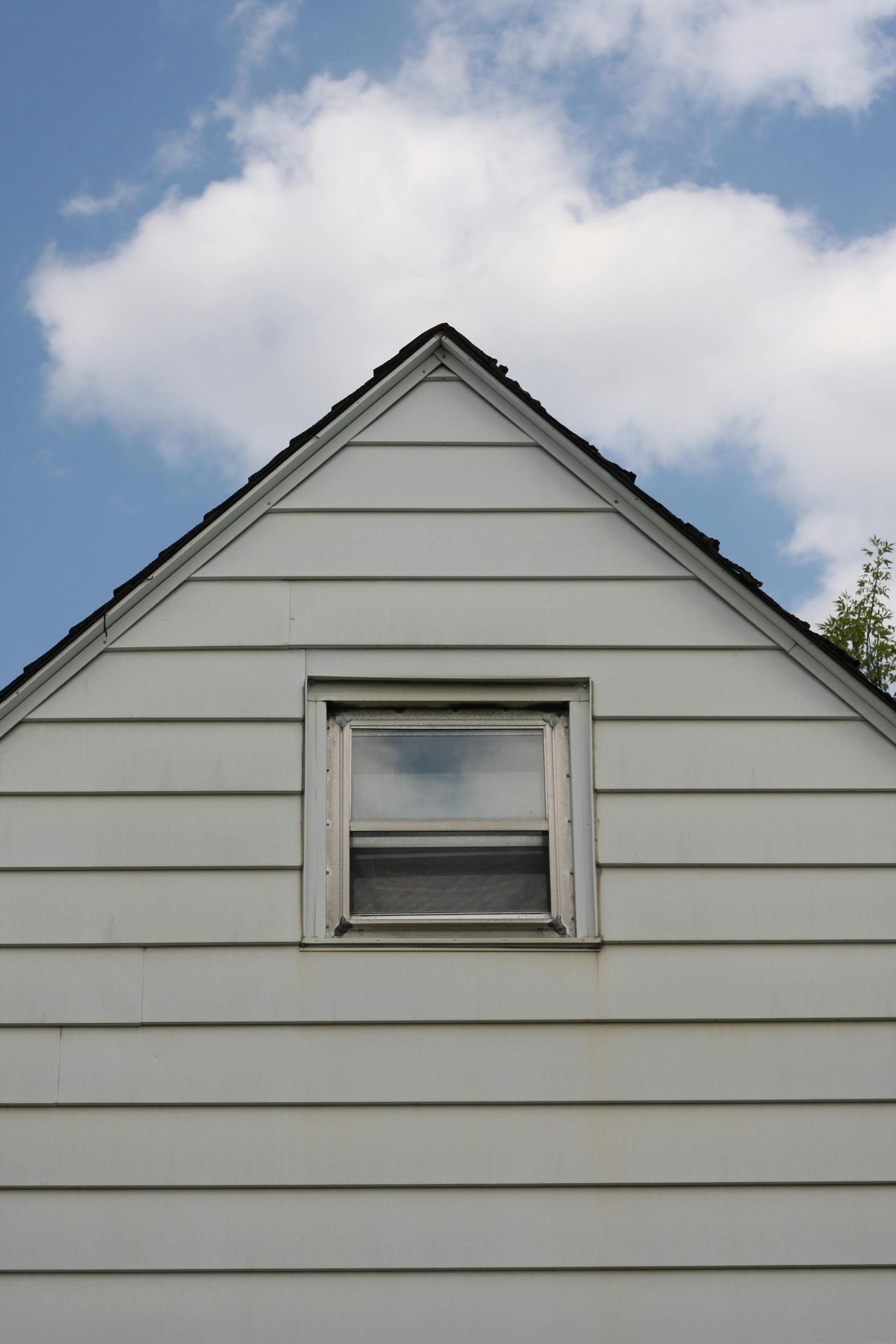 A white-sided gable of a house against a blue, cloudy sky, featuring a single centered square window.