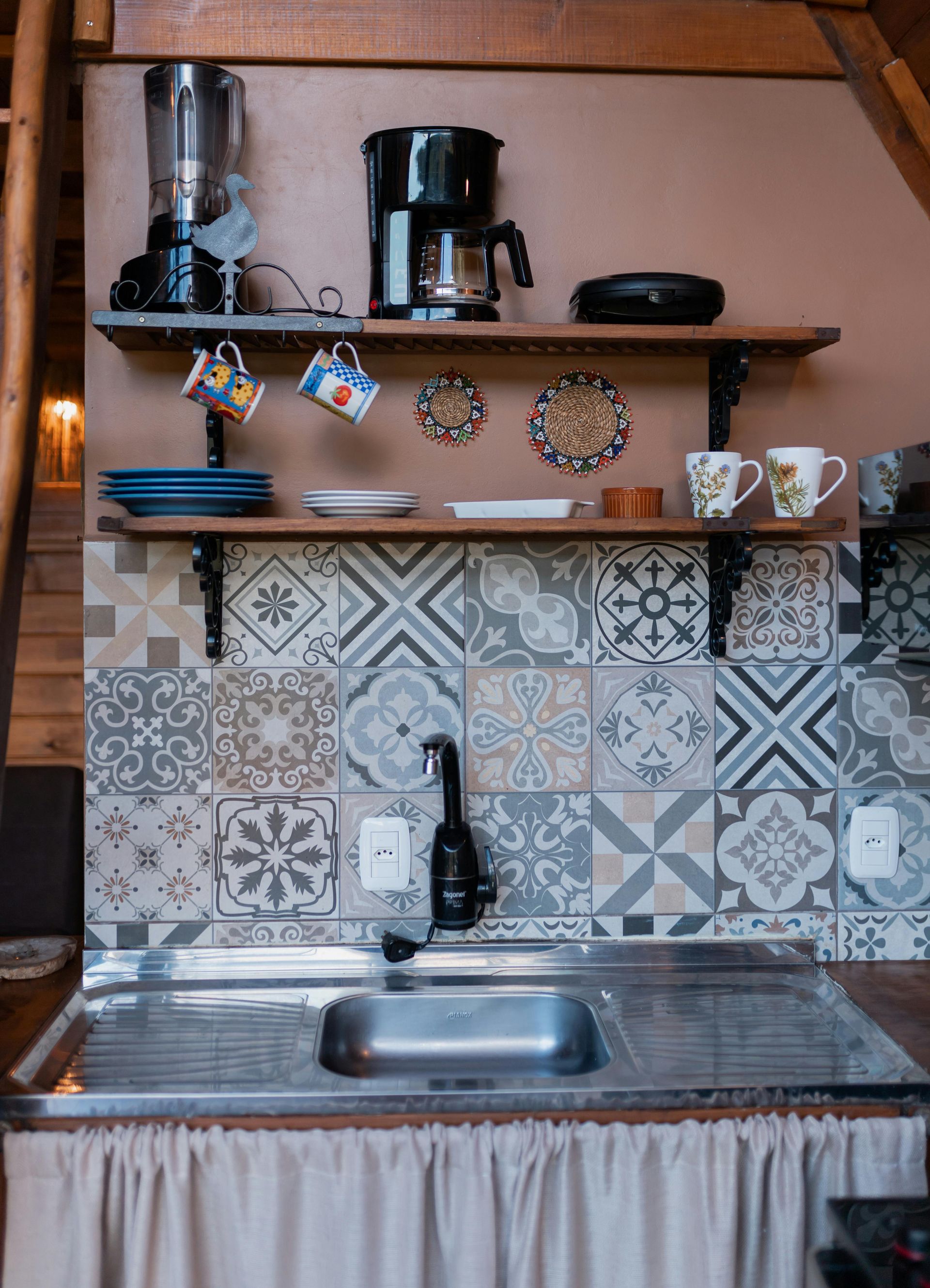 A kitchen sink area with patterned backsplash tiles, two wooden shelves holding mugs, a blender, and a coffee maker.