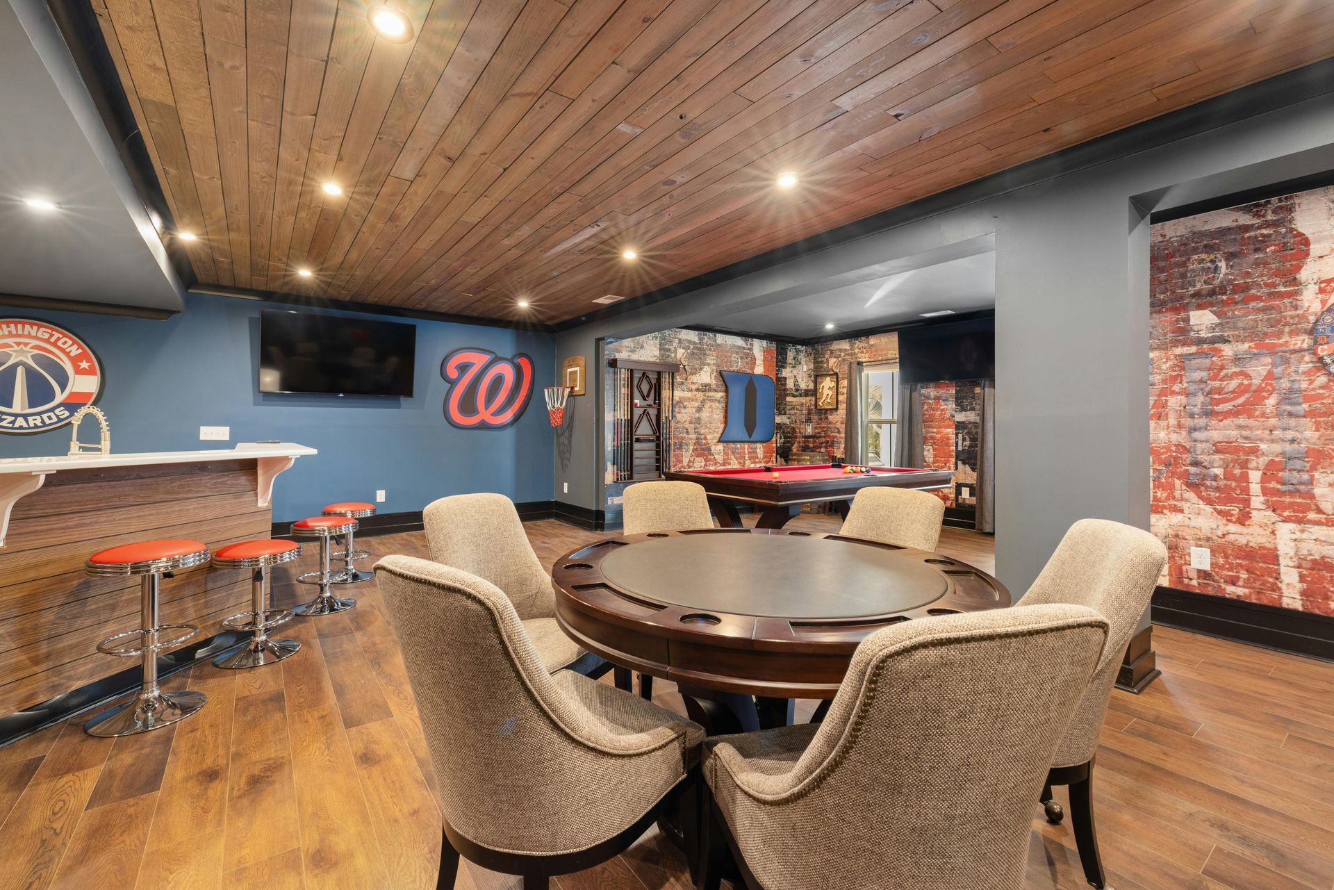 A finished basement featuring a circular poker table, a bar with orange stools, a pool table, and sports team logos.