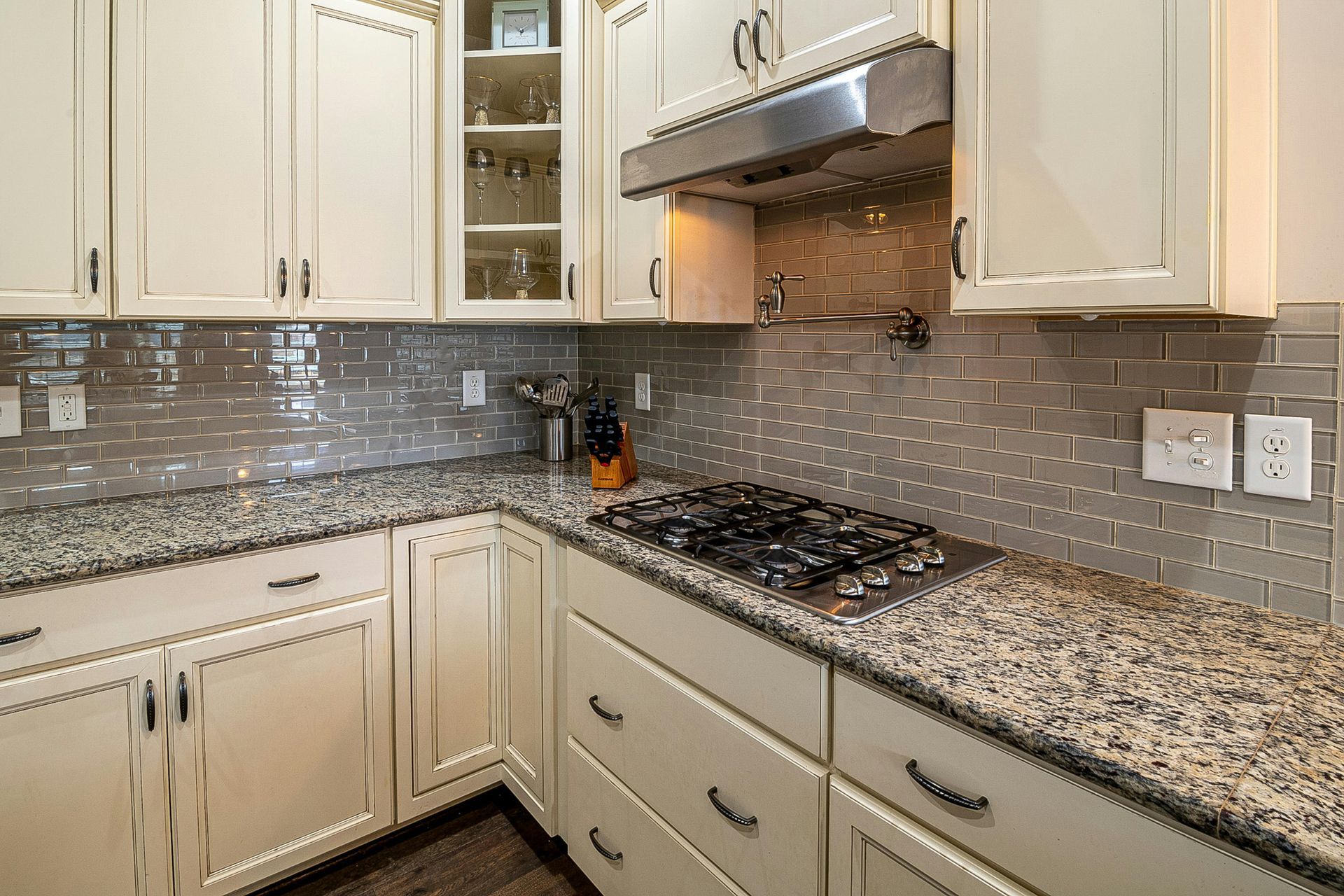 A kitchen with cream cabinets, speckled granite countertops, a stainless steel stove, and a grey subway tile backsplash.