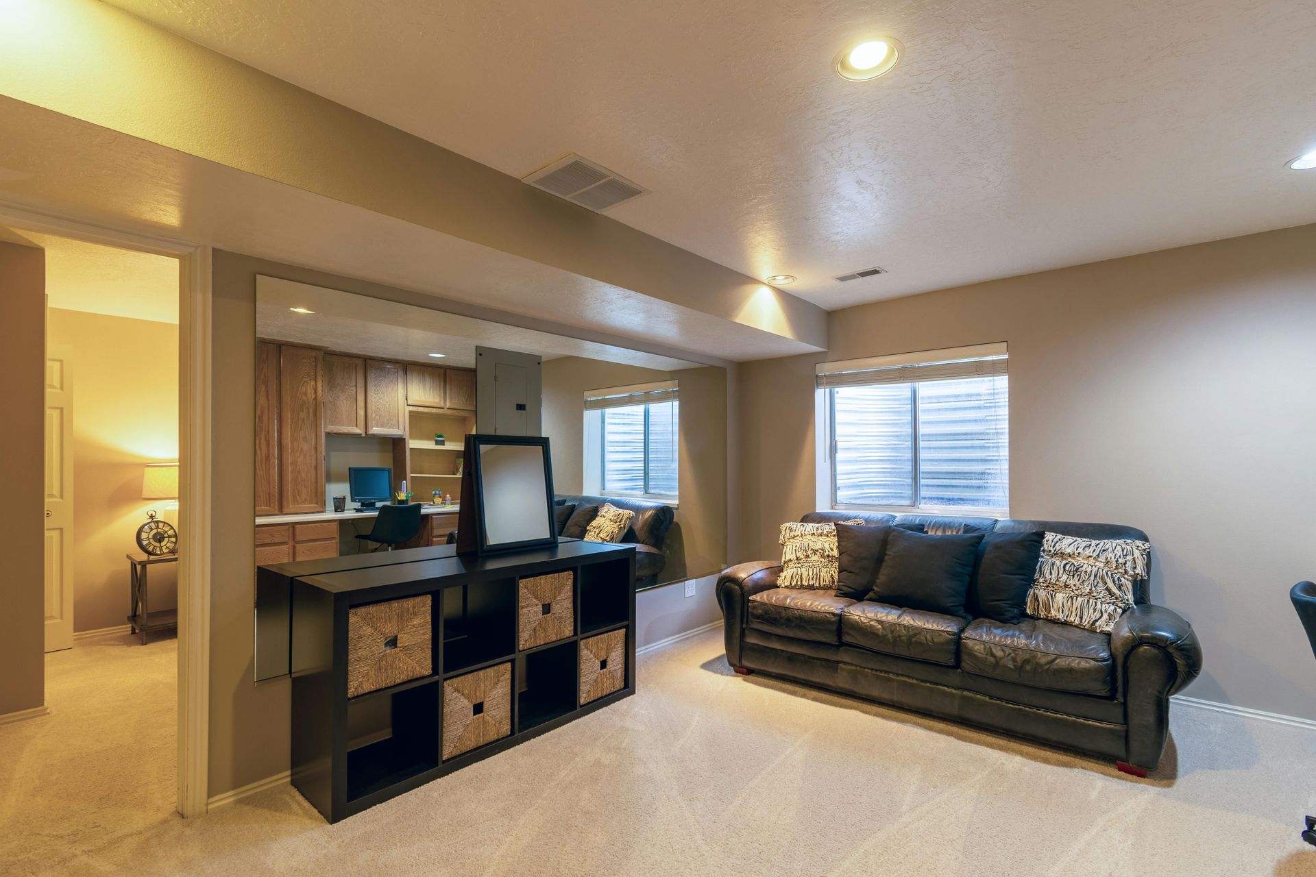 A carpeted basement with a black couch, a storage cube shelf, and an adjacent office area with wooden cabinets.