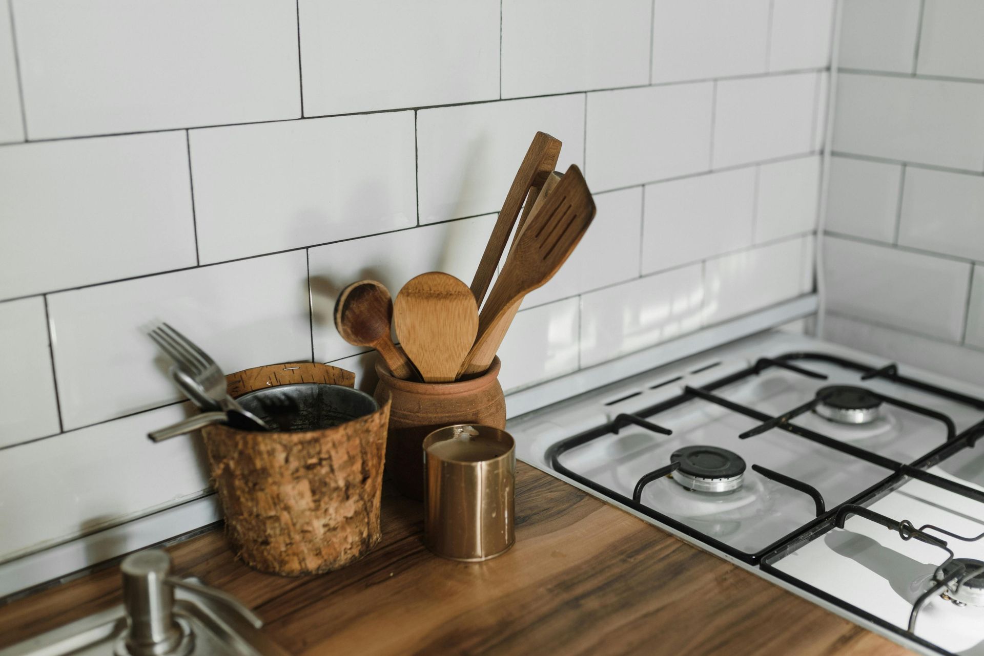Wooden and metal kitchen utensils in containers on a wooden countertop next to a stovetop against a white tiled wall.