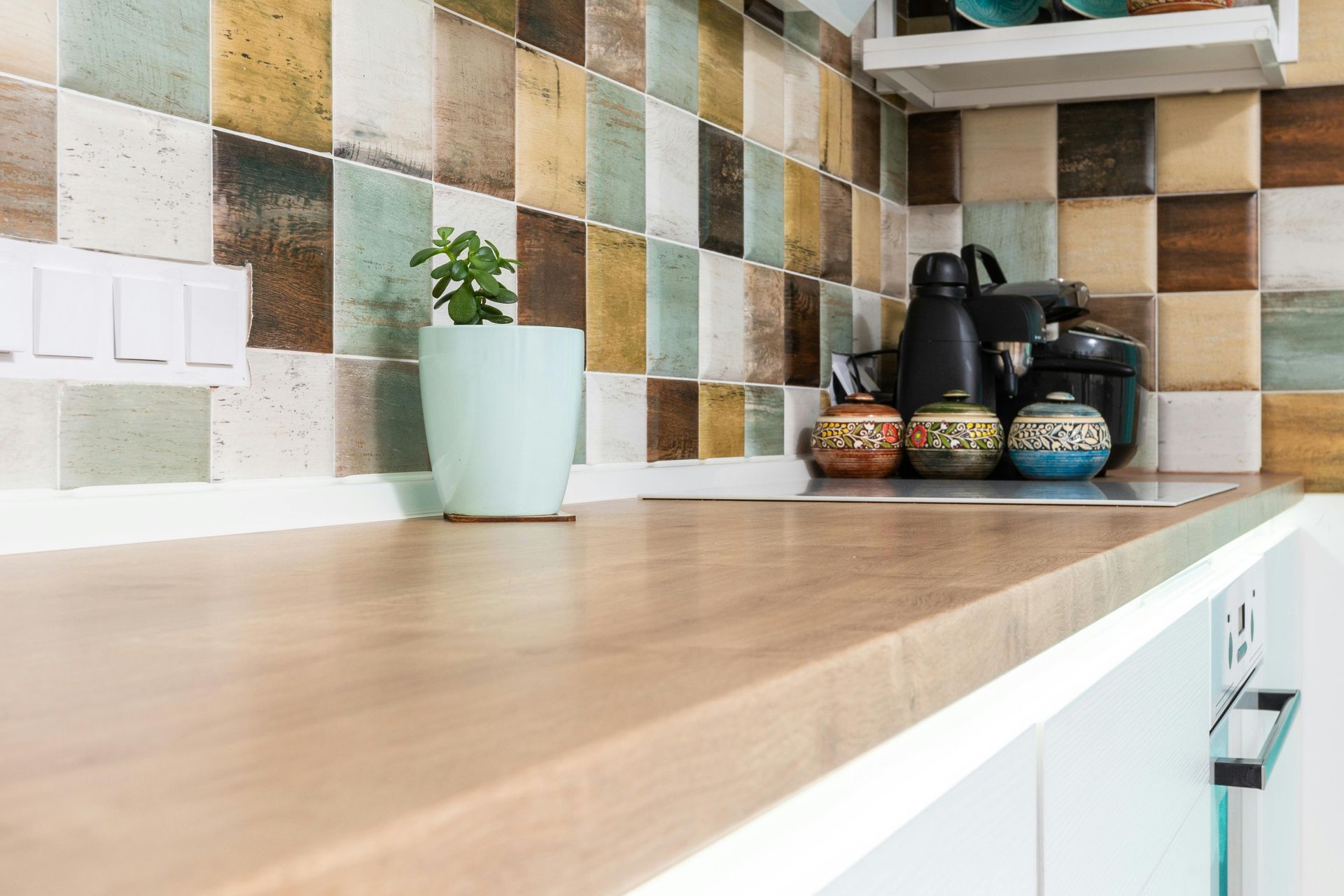 A light wood kitchen countertop next to a multicolored tiled backsplash with a small potted plant and coffee appliances.