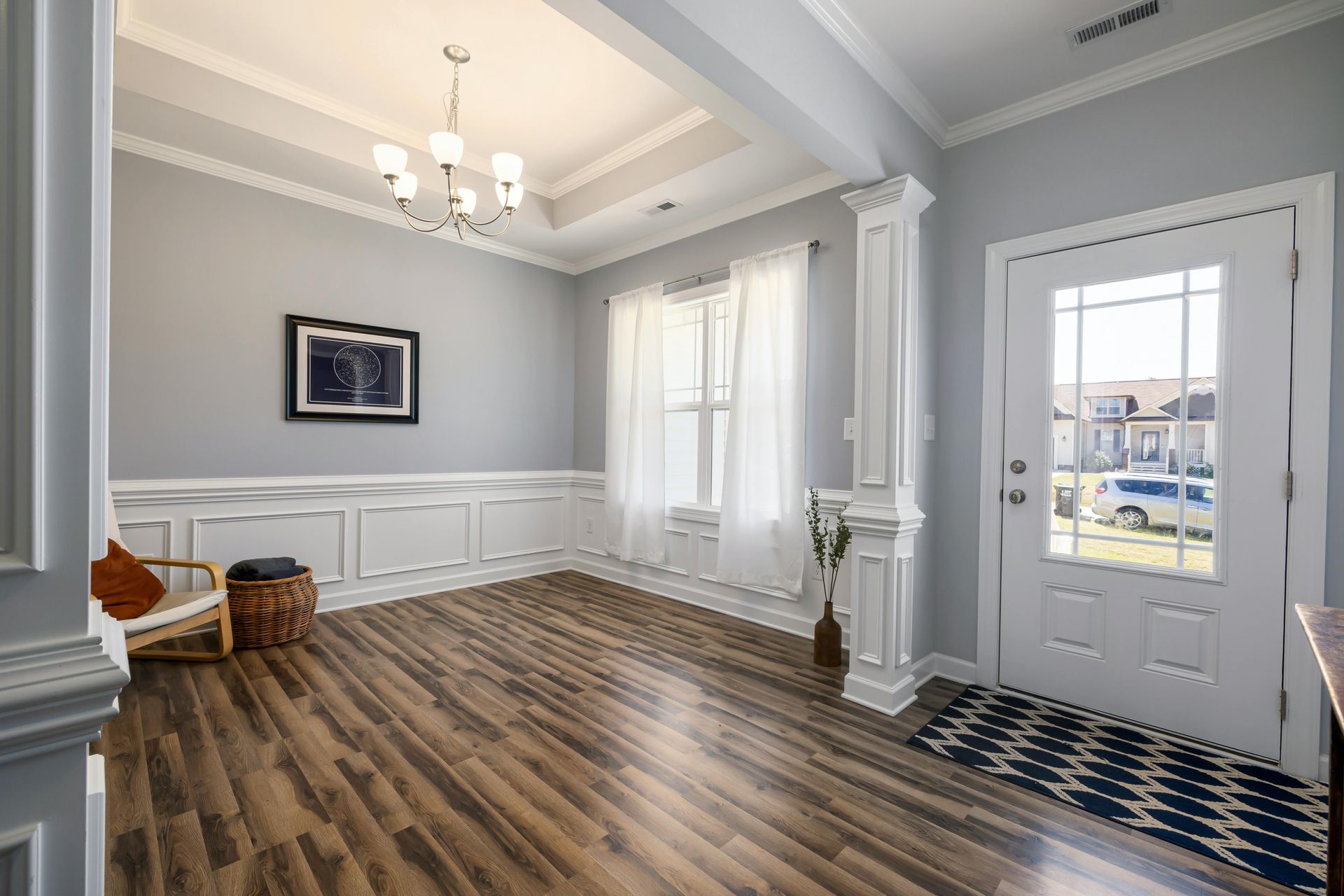 Bright foyer with wood flooring, white wainscoting, a chandelier, a chair, and a white front door with a glass panel.