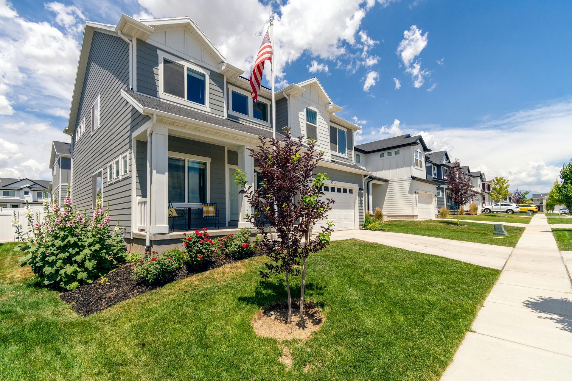 A modern two-story gray and white house with an American flag, green lawn, and sidewalk under a cloudy blue sky.