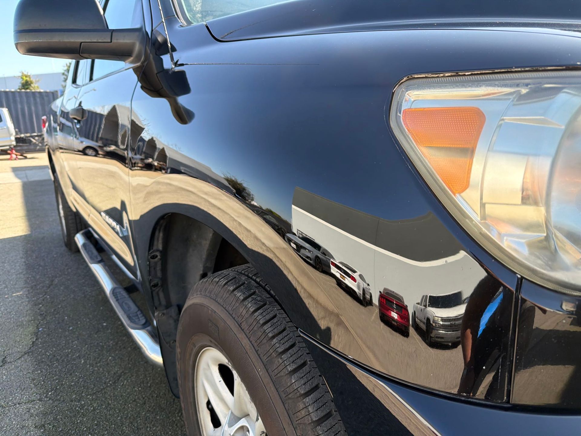 Black truck's front quarter panel reflecting the surrounding environment, including vehicles and buildings.