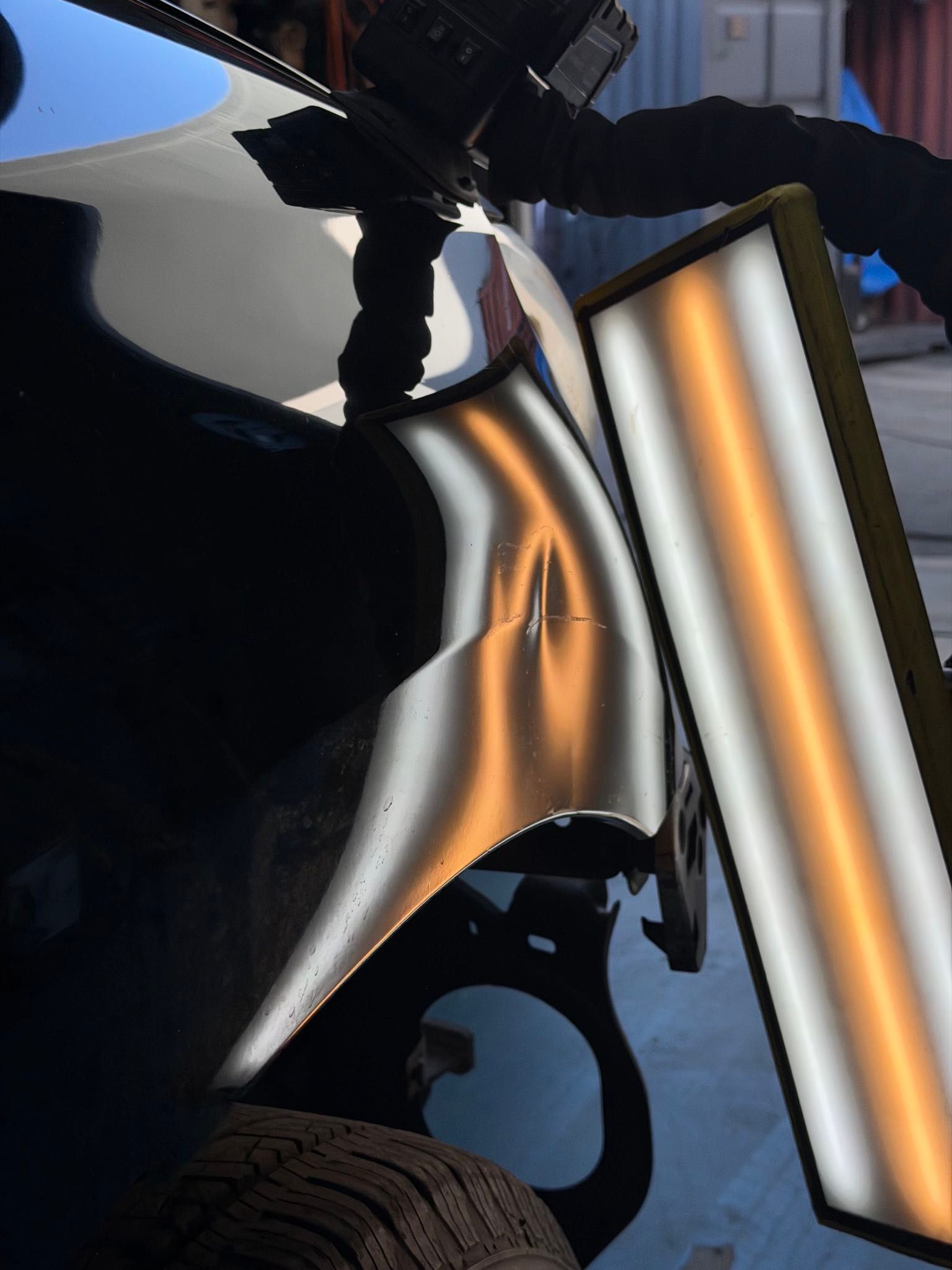 Car fender dent being repaired with specialized tools, reflecting a light panel.