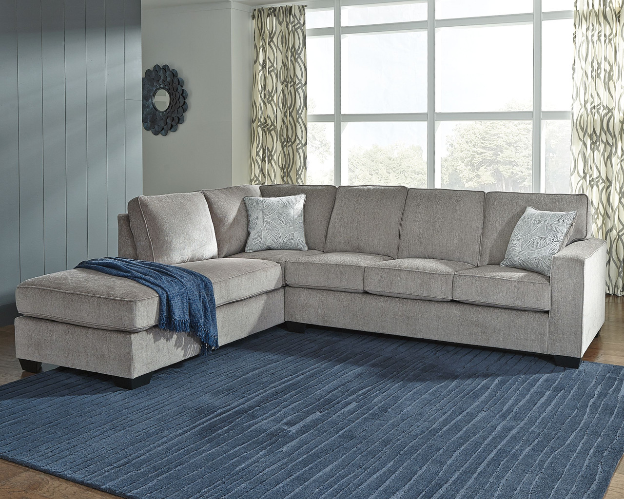L-shaped gray sofa with blue pillows and blanket on a blue rug in front of a window.