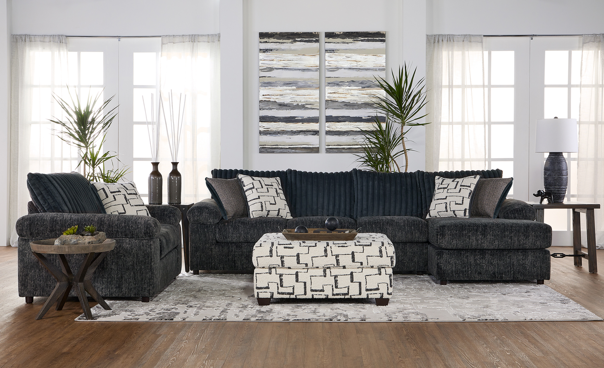 Living room furniture set: dark gray sectional sofa, armchair, ottoman, and wall art in front of windows.