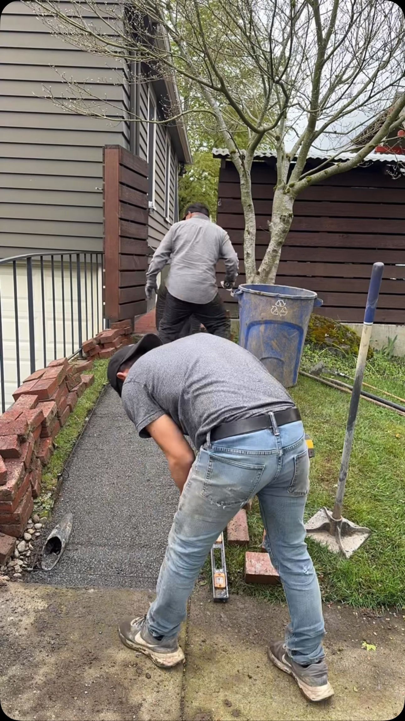 Two workers laying gravel path next to a building, one digging, one carrying supplies.