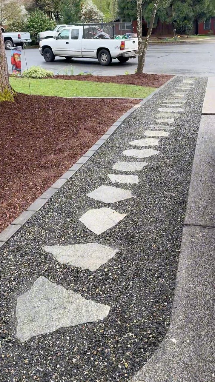 Stone path leads through dark gravel, bordered by mulch and concrete, with a truck in the background.