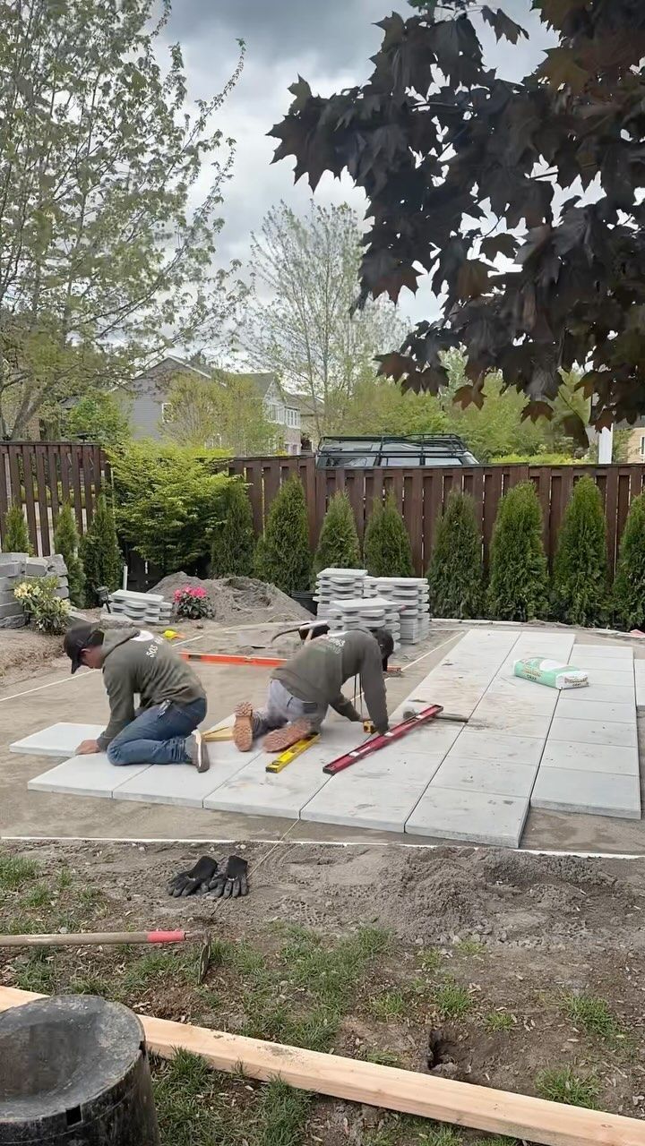 Two workers laying paving stones in a backyard. Green shirts, kneeling, measuring. Cloudy day.