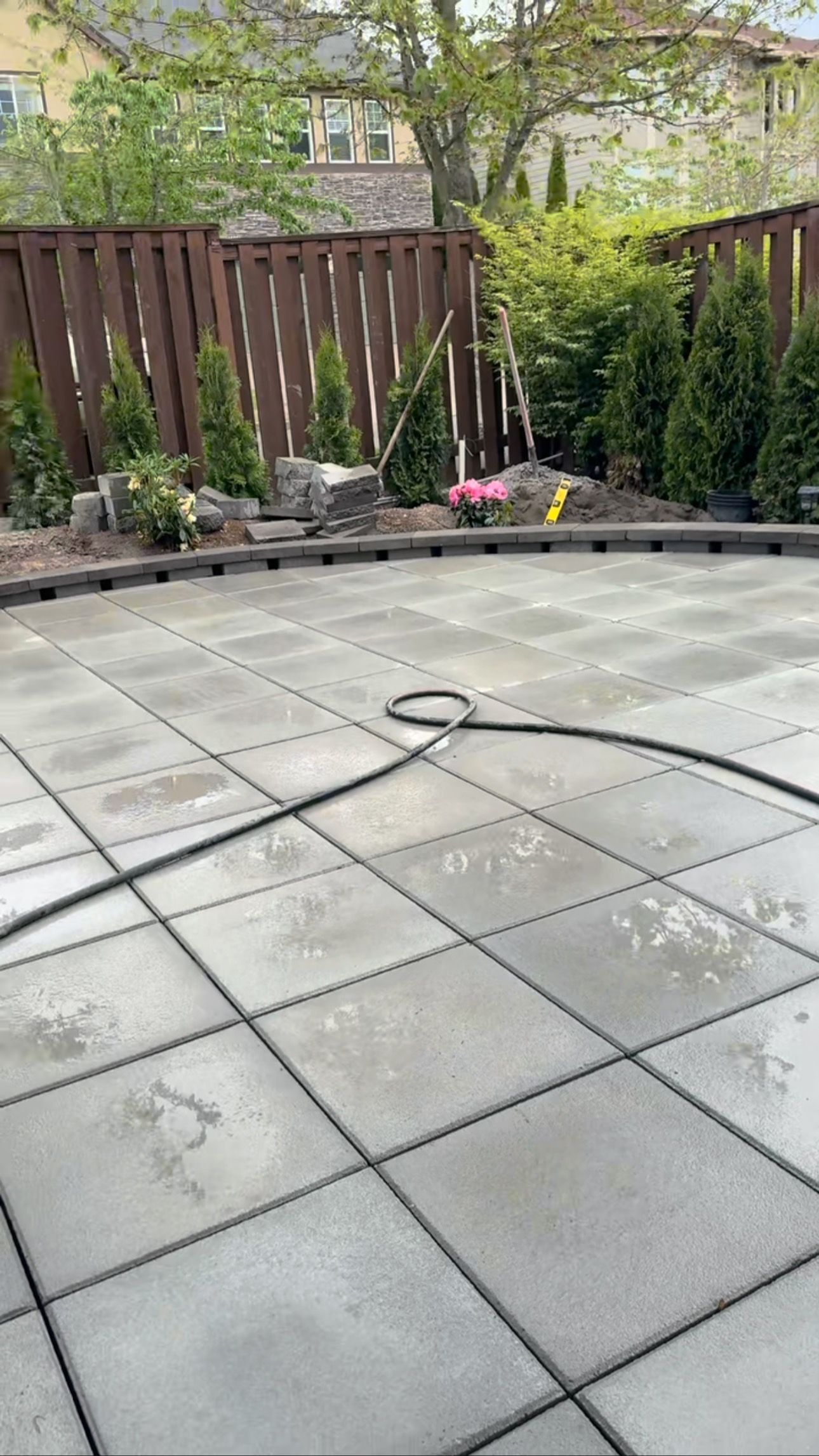Patio with square concrete tiles and a hose in front of a brown fence and greenery.