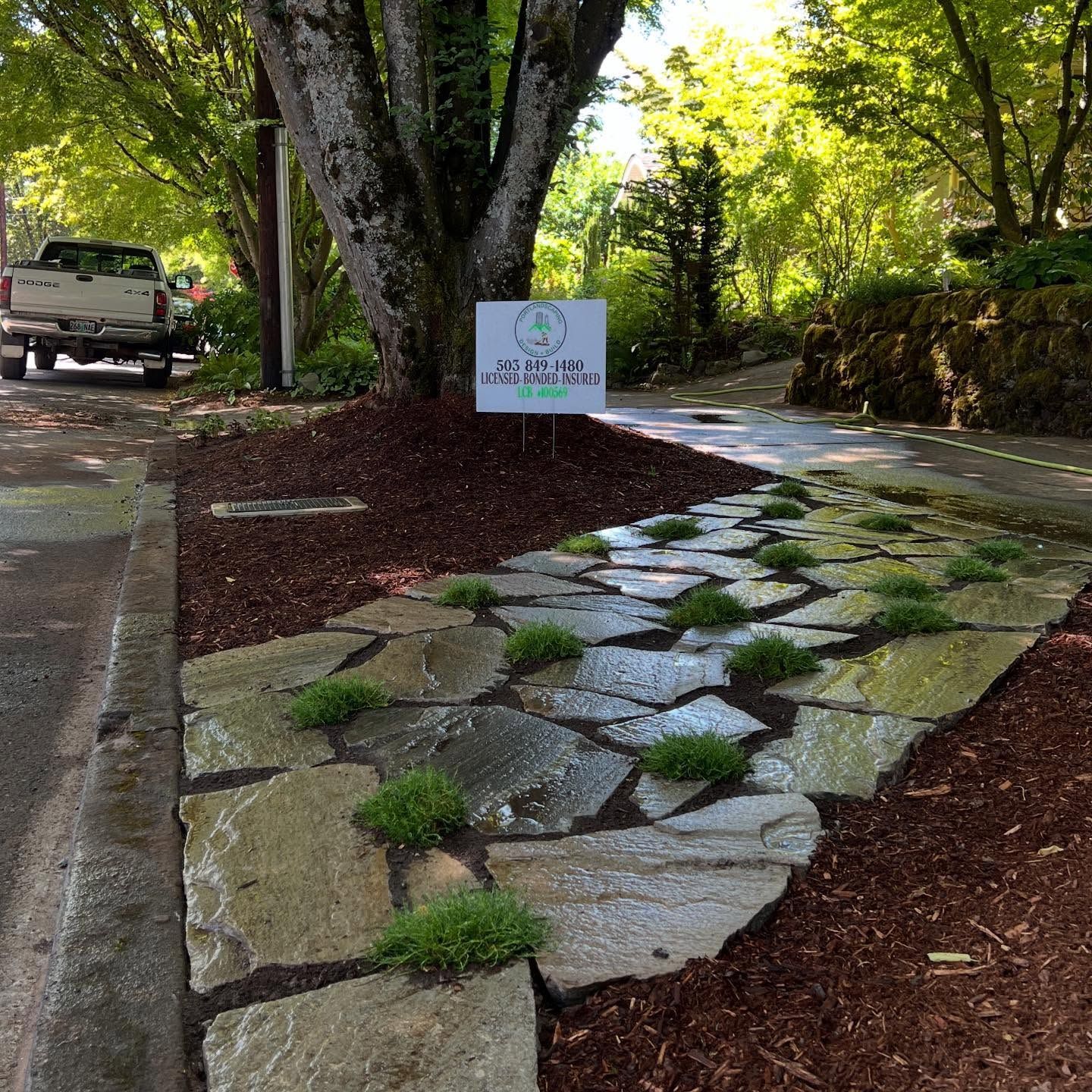 Flagstone pathway with green plants, next to a driveway with a truck, and sign under a large tree.