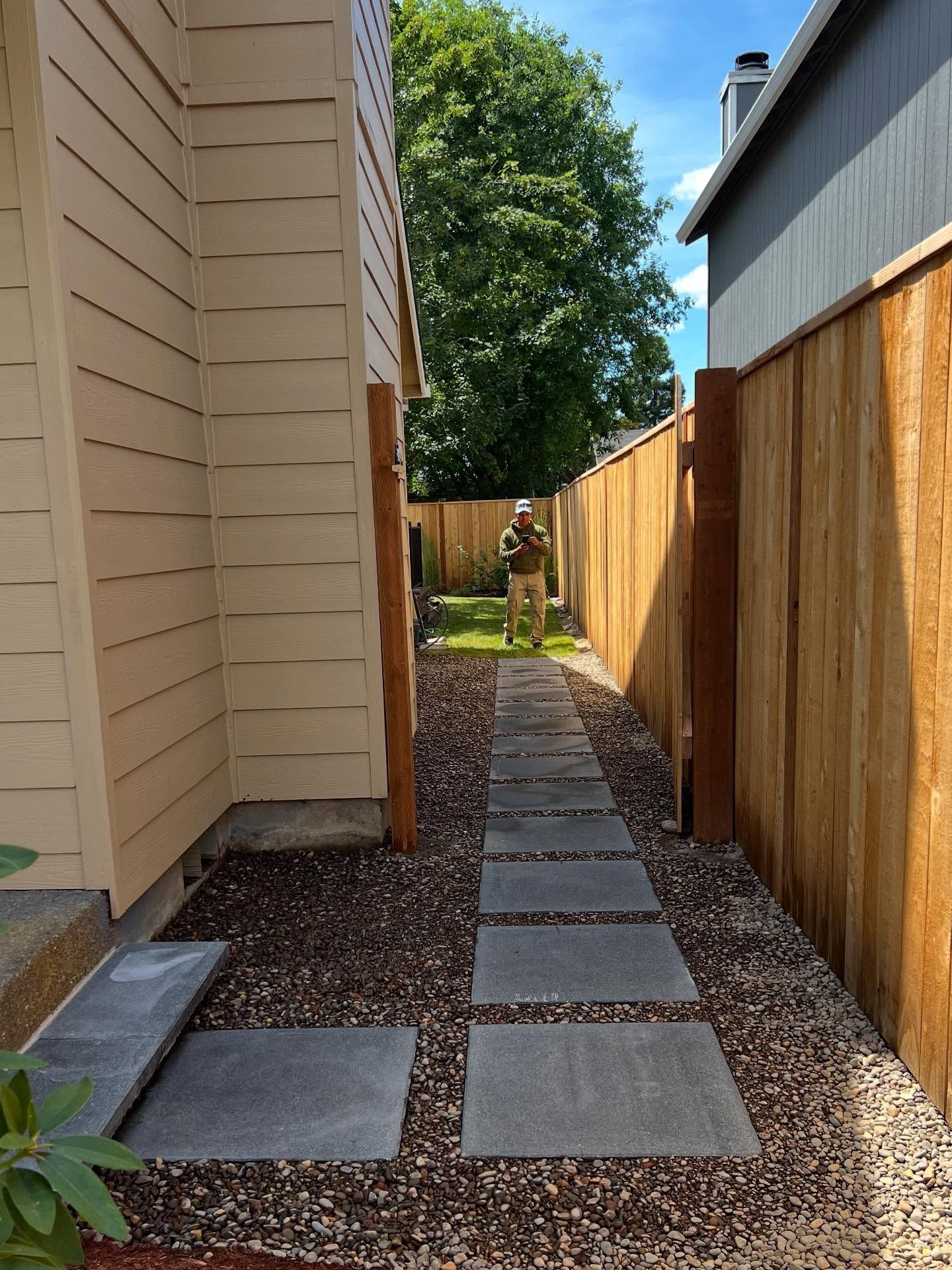 Path between tan house and wooden fence. Person standing in distance.