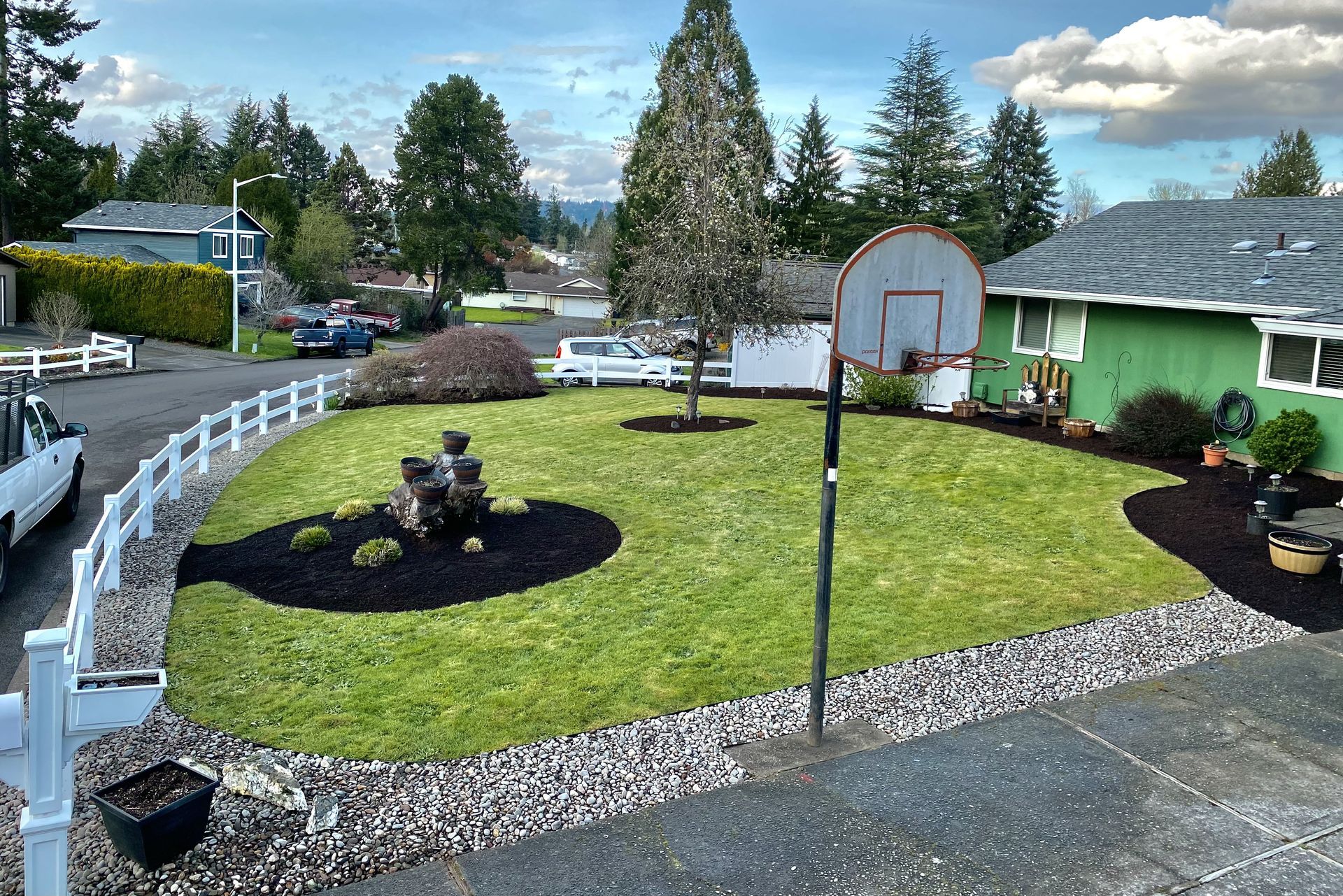 A well-manicured green lawn with a white fence, basketball hoop, and a green house in a suburban neighborhood.