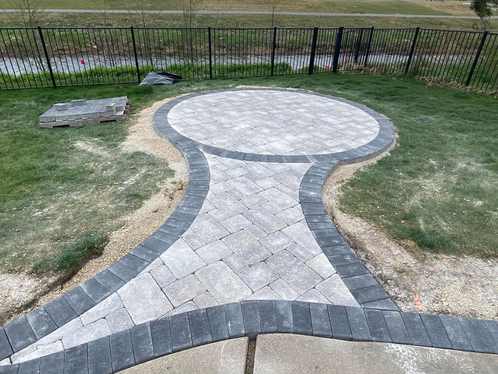 A gray brick patio and walkway lead to a circular patio area, edged in black brick, on a grassy lawn. A gray brick patio and walkway lead to a circular patio area, edged in black brick, on a grassy lawn.