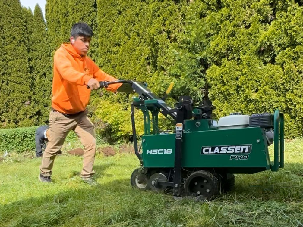 Young man in orange hoodie operating a green Classen sod cutter on grass; backyard setting. Young man in orange hoodie operating a green Classen sod cutter on grass; backyard setting.