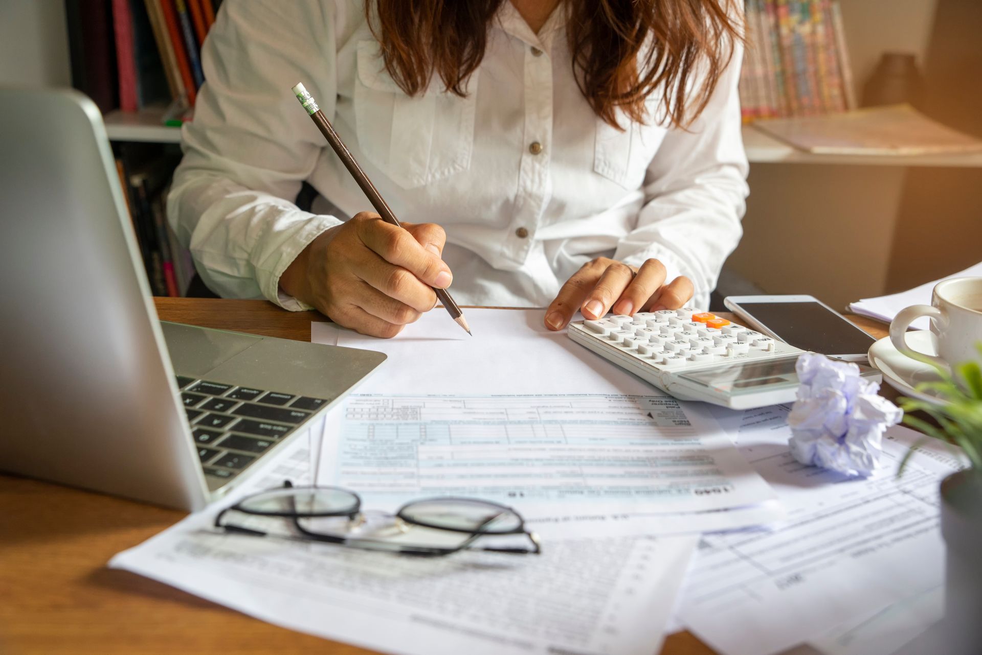 A person in a white shirt writes on financial forms at a desk with a laptop, calculator, and glasses.